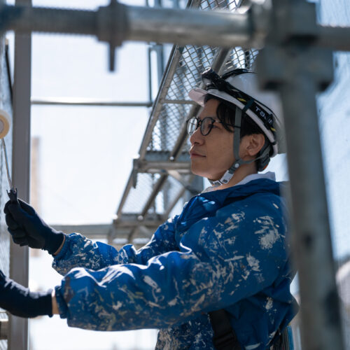 Construction worker in a helmet and gloves paints a wall with a roller on scaffolding nearby.