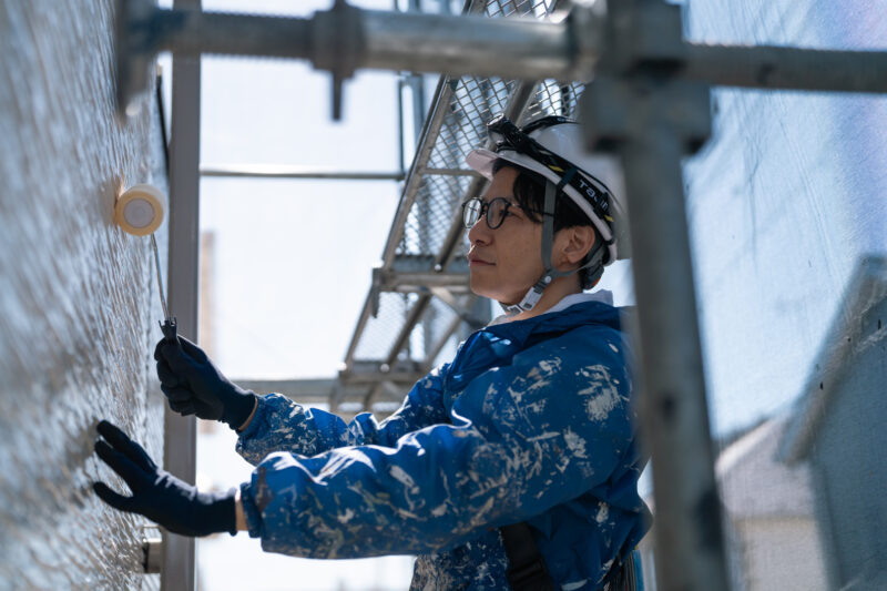 Construction worker in a helmet and gloves paints a wall with a roller on scaffolding nearby.