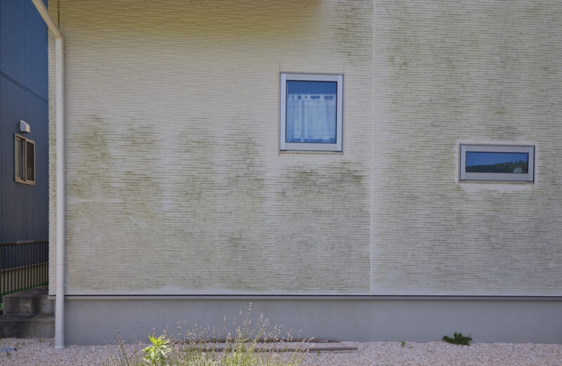 pixta_106187908_M - 堀岡美装店 Beige textured building wall with two rectangular windows, left side blue panel, and gravel foreground with sparse plants.