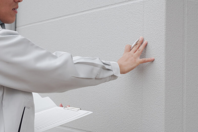 pixta_112184768_M - 堀岡美装店 Person in a light gray uniform places a hand on a textured wall while holding a clipboard nearby, suggesting inspection or assessment.