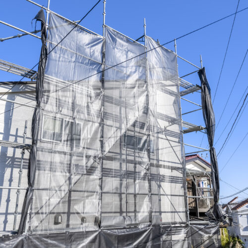 Construction site around a house with metal scaffolding and gray safety netting under a clear blue sky