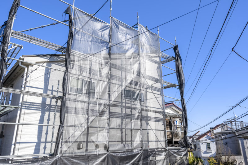 pixta_139402963_L - 堀岡美装店 Construction site around a house with metal scaffolding and gray safety netting under a clear blue sky