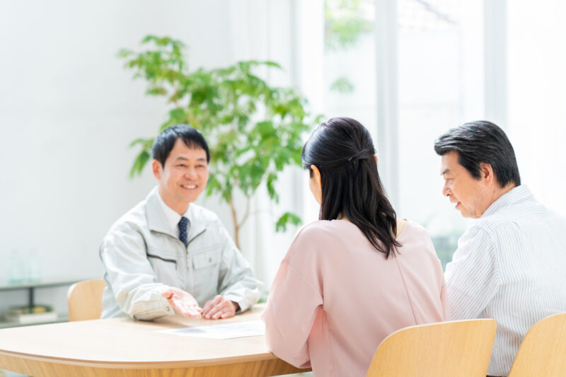 pixta_77630852_M - 堀岡美装店 Three colleagues in a bright office seated around a curved wooden table, chatting by a window with a plant in the background.