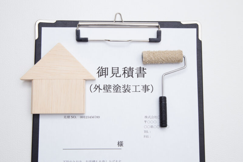 pixta_90671683_M - 堀岡美装店 Clipboard with a Japanese quotation form (御見積書) and a small wooden house model plus a paint roller laid on top, symbolizing a home exterior painting quote.
